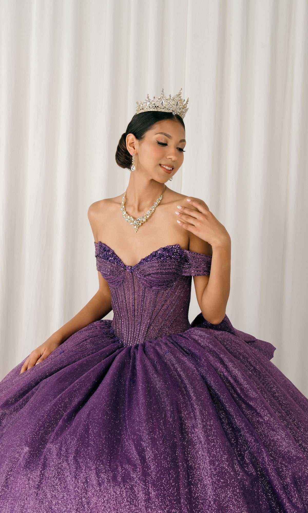A woman in a sparkly purple Quinceanera Dress 8460JQ By Cinderella Couture and tiara poses against a white curtain, accessorized with a necklace and earrings, ready for her special night in this stunning glitter gown.